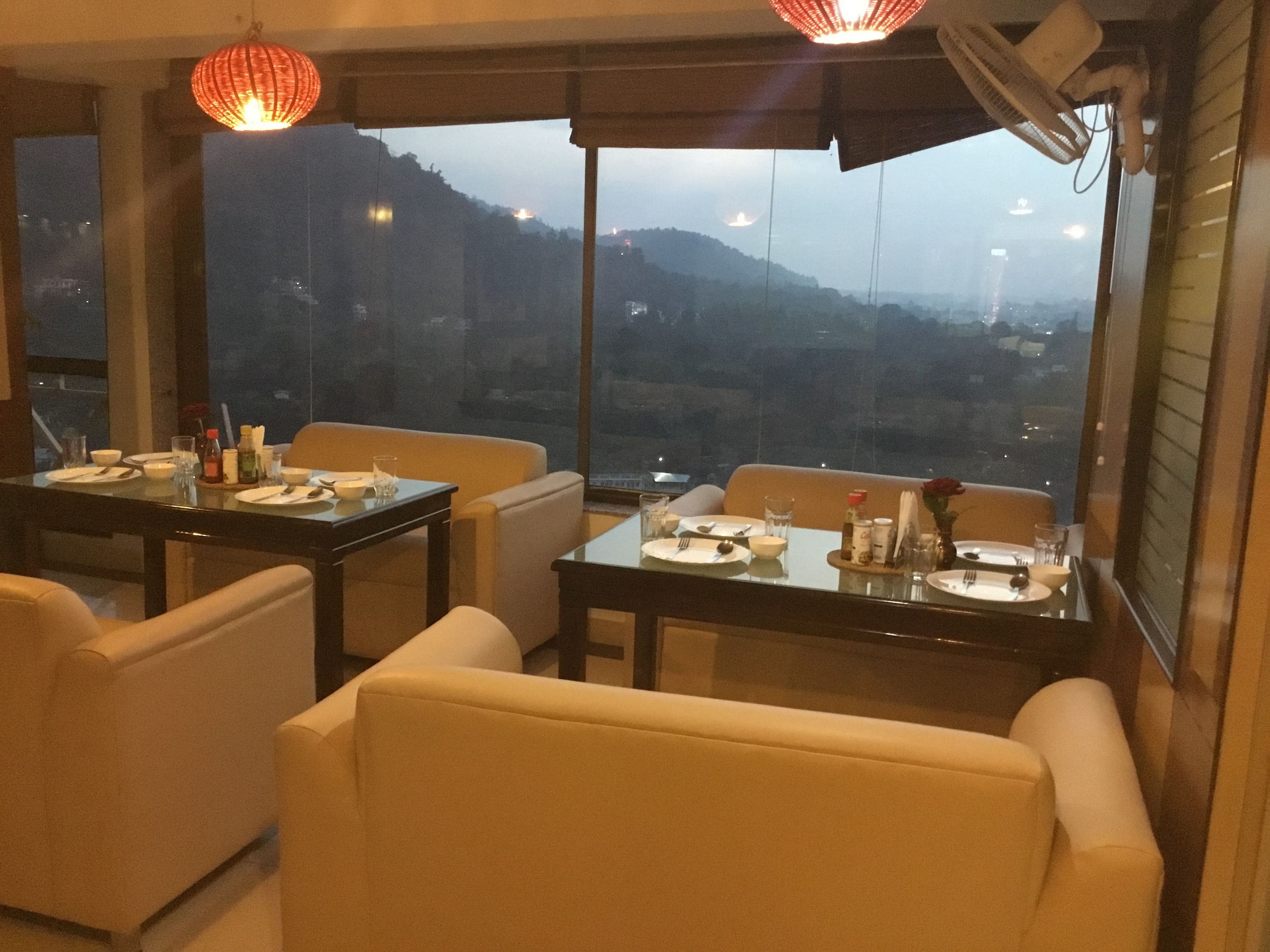 A restaurant with tables set for dining, large windows offering a view of the hills, and hanging lights.