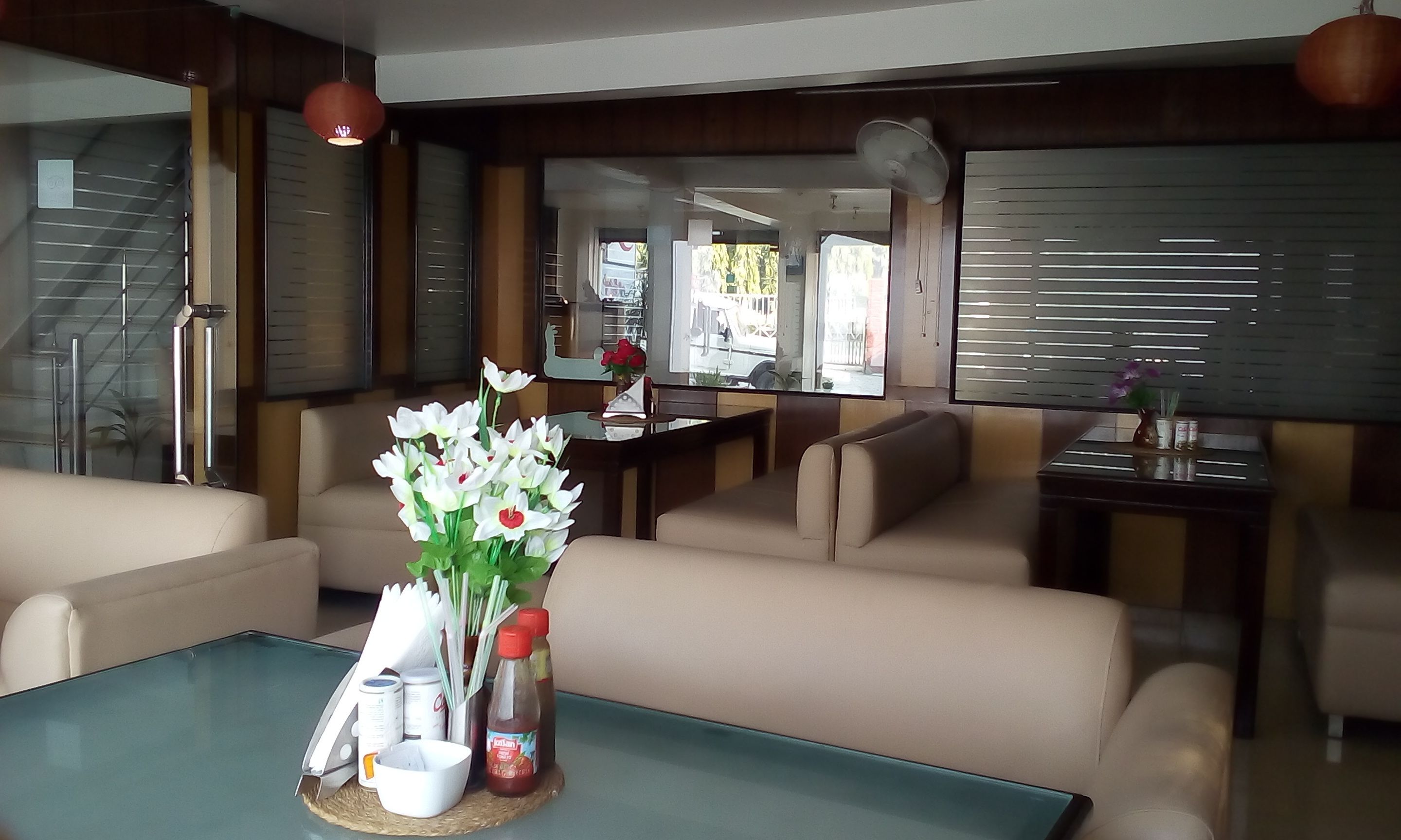 A modern hotel lobby with a glass coffee table, beige sofas, and a dining area with a view of the street.