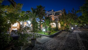 A courtyard with a cobblestone path, palm trees, and a building with arched windows. - Pantheon Hotel (Santorini)