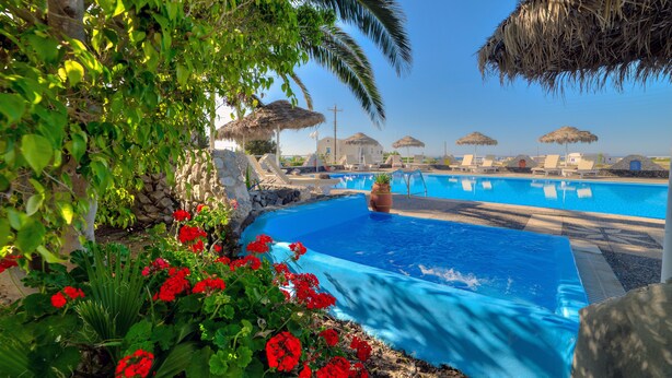 A swimming pool with a clear blue water feature, surrounded by lush greenery and red flowers. - Pantheon Hotel (Santorini)