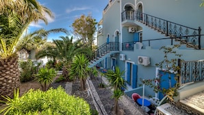 A multi-story building with a blue exterior, a staircase with black railings, and a garden with palm trees and shrubs. - Pantheon Hotel (Santorini)