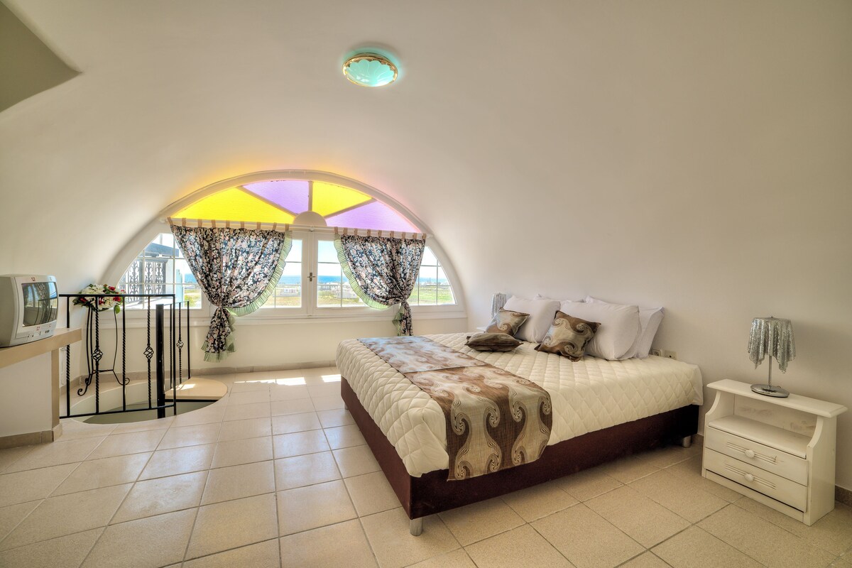 A bedroom with a large bed, a bedside table, a small table with a lamp, and a window with curtains.