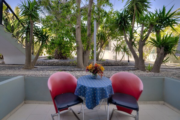 A small outdoor patio with a table and two chairs, surrounded by trees and a stone pathway. - Pantheon Hotel (Santorini)