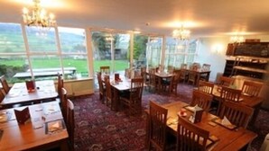 A dining area with wooden tables and chairs, large windows offering a view of the outdoors, and a chandelier hanging from the ceiling.