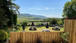 A patio area with wooden tables and chairs, surrounded by a wooden fence, and overlooking a scenic landscape with hills and trees.