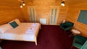 A cozy wooden cabin room with a bed, a green chair, a small table, and a radiator.