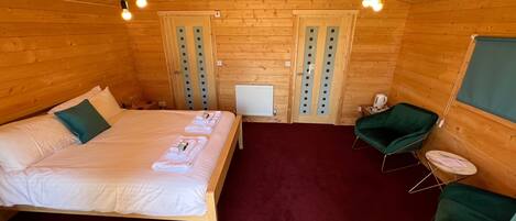 A cozy wooden cabin room with a bed, a green chair, a small table, and a radiator.