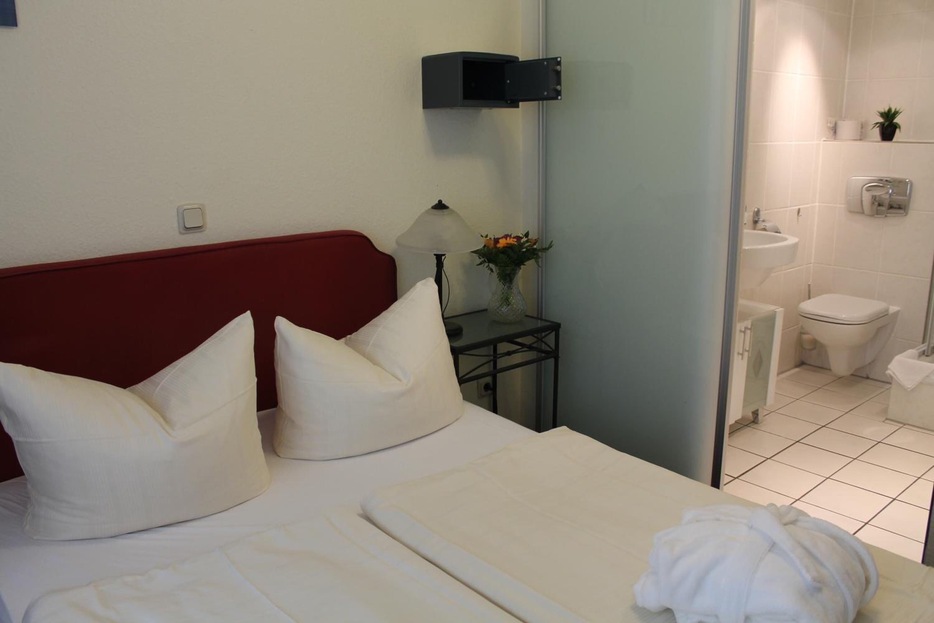 A hotel room with a bed, pillows, a nightstand with a lamp and vase, a red headboard, and a bathroom visible through an open door.