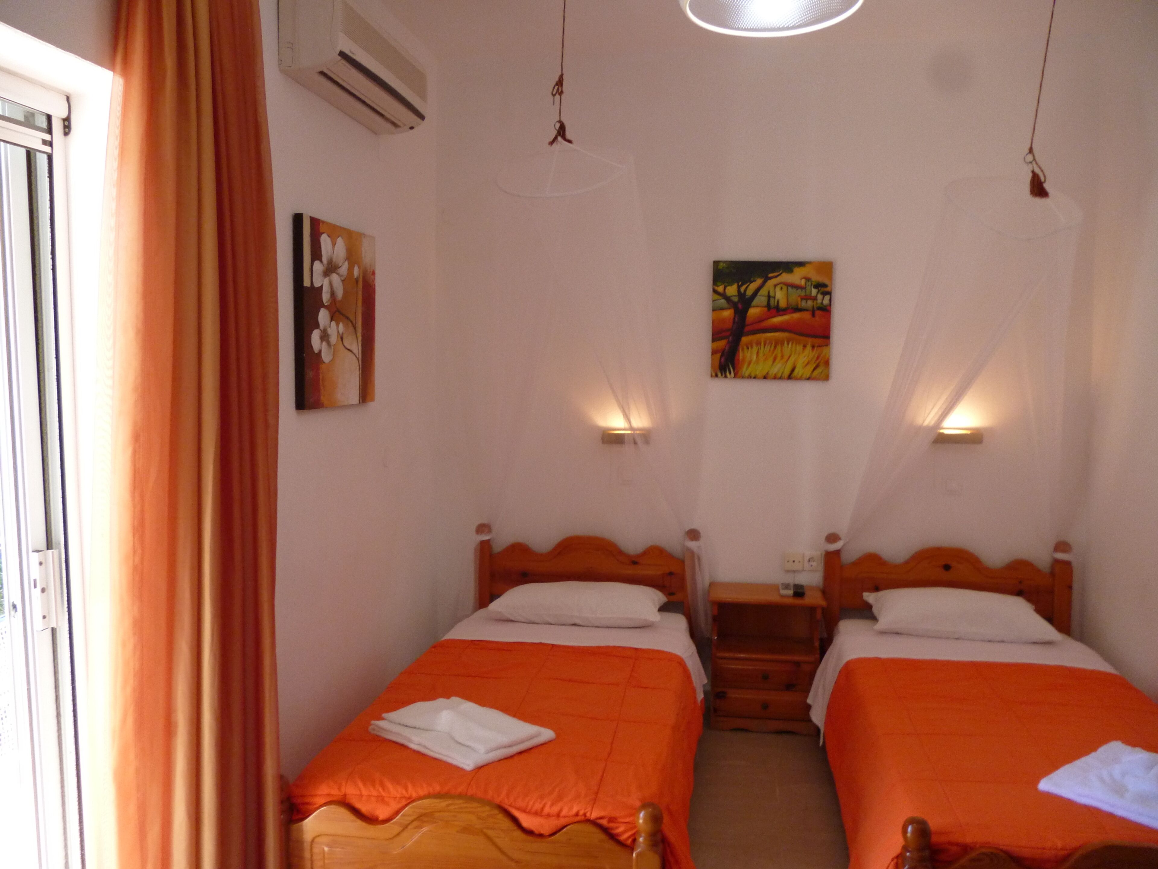 A room with two beds, each with an orange bedspread and white pillows. There are two wall-mounted lamps, a painting on the wall, and an air conditioning unit.