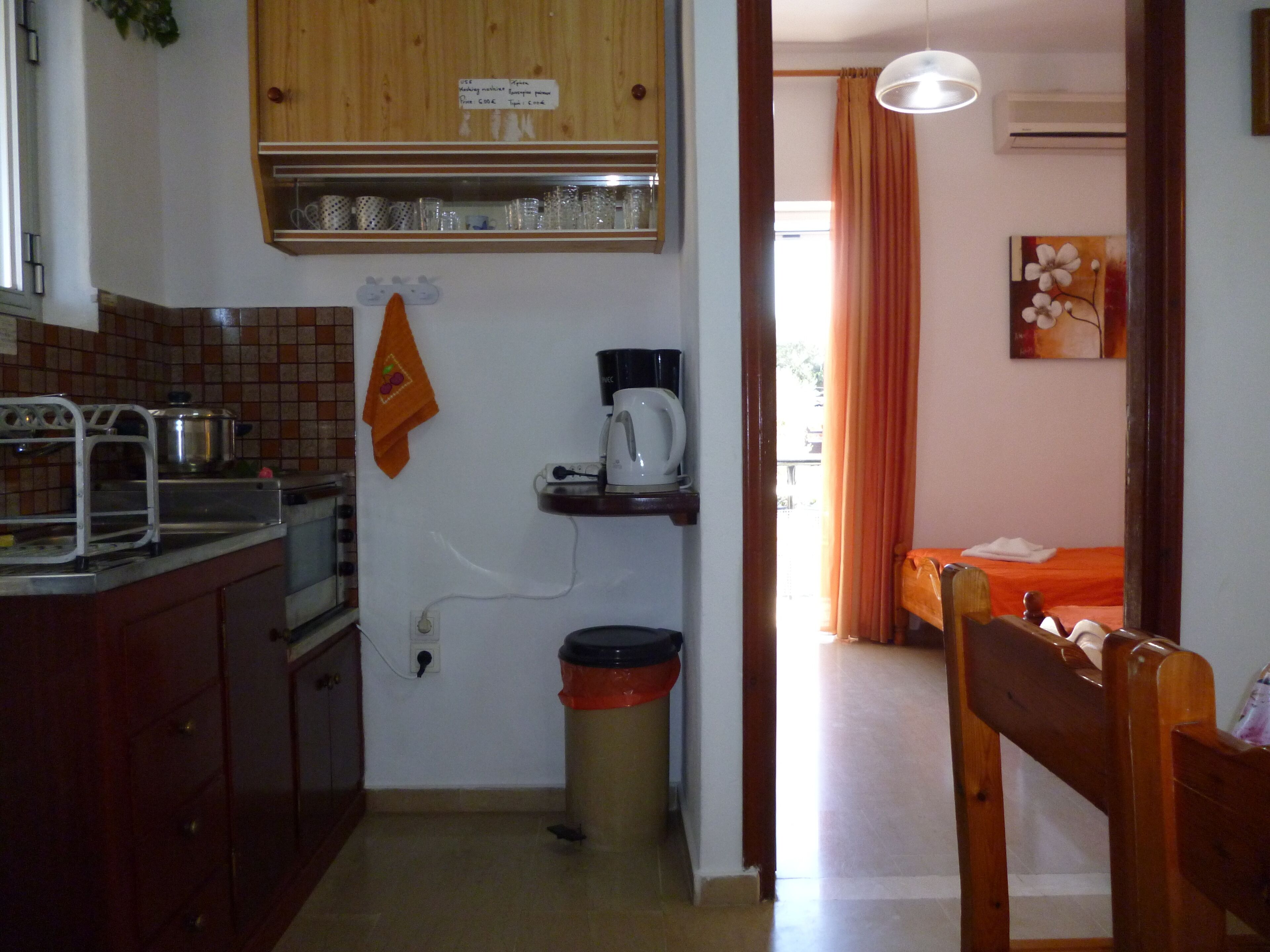 Une petite cuisine équipée d’une cuisinière, d’un évier et d’une machine à café posée sur une étagère. On aperçoit une poubelle et une chaise en bois au premier plan.