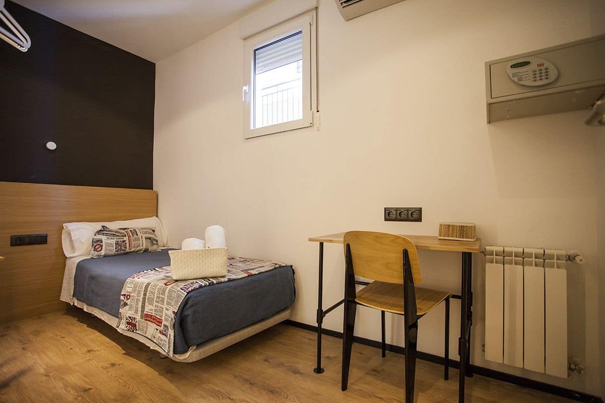 Quarto pequeno e bem organizado, com uma cama, uma cadeira, uma mesa e uma janela.