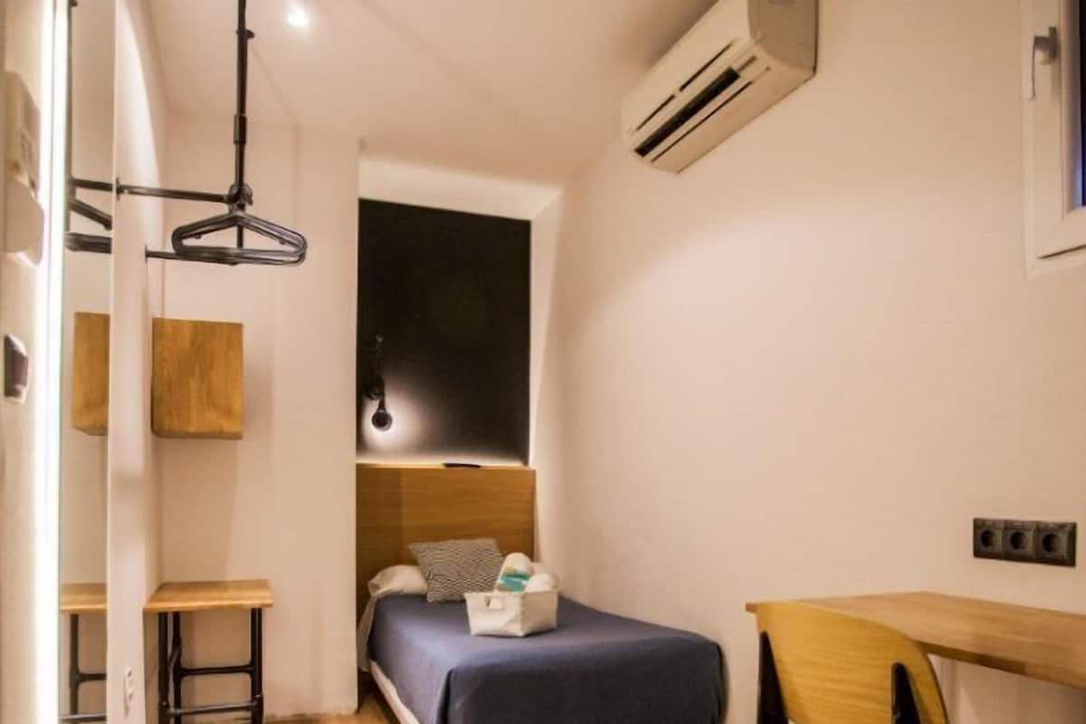 Quarto compacto com cama, escrivaninha, cadeira, luminária pendente e ar condicionado.
