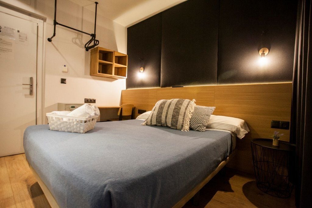 A modern hotel room with a large bed, a wooden headboard, a small bedside table, and a wall-mounted shelf.