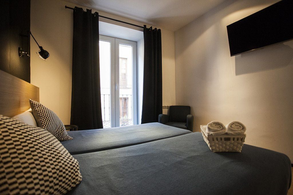 Ein Hotelzimmer mit zwei Betten, einem Sessel, einem an der Wand befestigten Fernseher und einem Fenster mit Vorhängen.