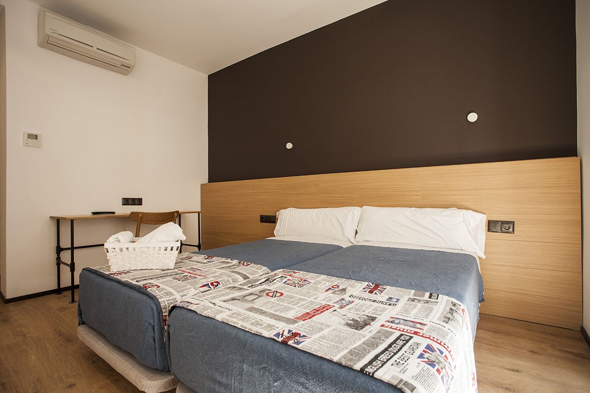 Een hotelkamer met een houten hoofdeinde, een bed met een deken met een krantenprint, een nachtkastje met een mand, en een klein bureau met een stoel.