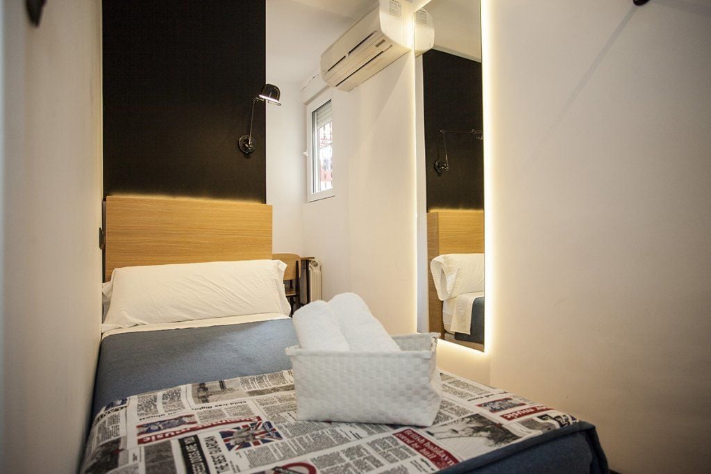 A hotel room with a bed, a bedside table with a newspaper, a wall-mounted air conditioner, and a window.
