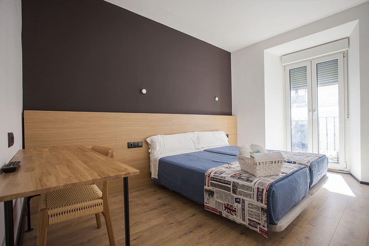 Quarto moderno com cabeceira de madeira, uma cama de solteiro com colcha azul, uma mesa de madeira com cadeira e uma janela grande com persianas.