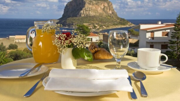A table set with a pitcher of orange juice, a glass of water, a cup, a plate, a napkin, a spoon, and a croissant, with a scenic view of a mountain and sea in the background. - Hotel Panorama (Monemvasia)