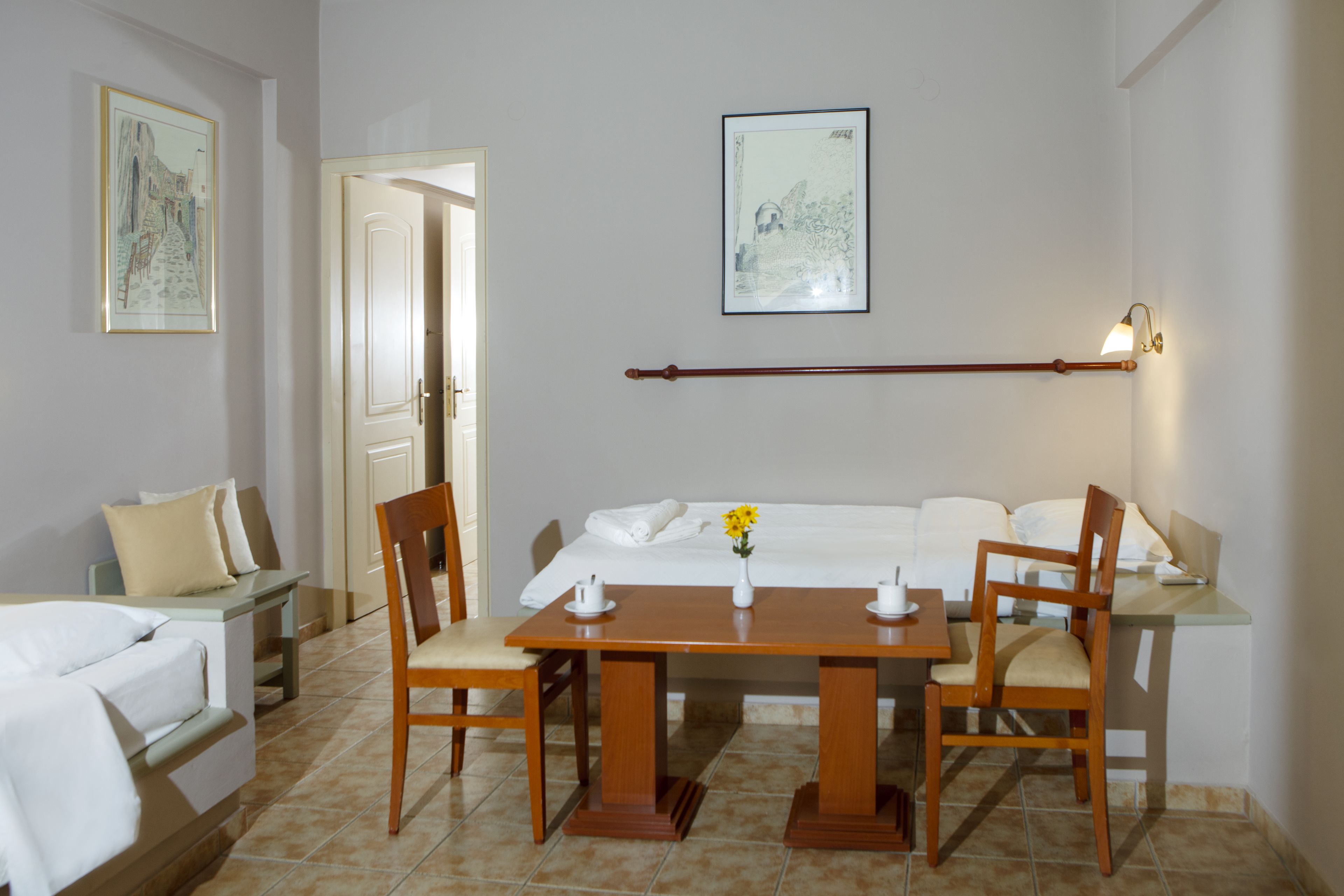 Una habitación de hotel con una cama, una mesa de madera con sillas, una mesita con un florero y dos cuadros en la pared.