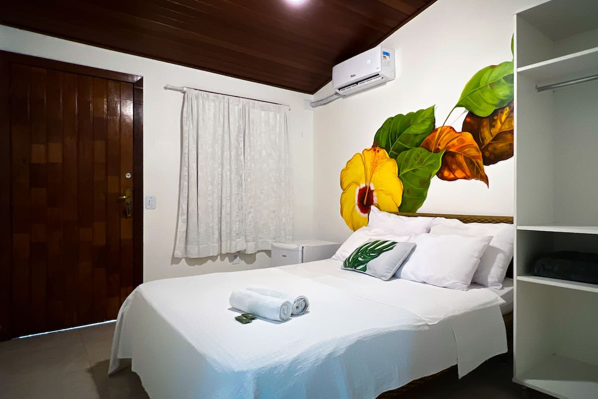 A hotel room with a bed, a white curtain, an air conditioning unit, and a shelf with a plant.