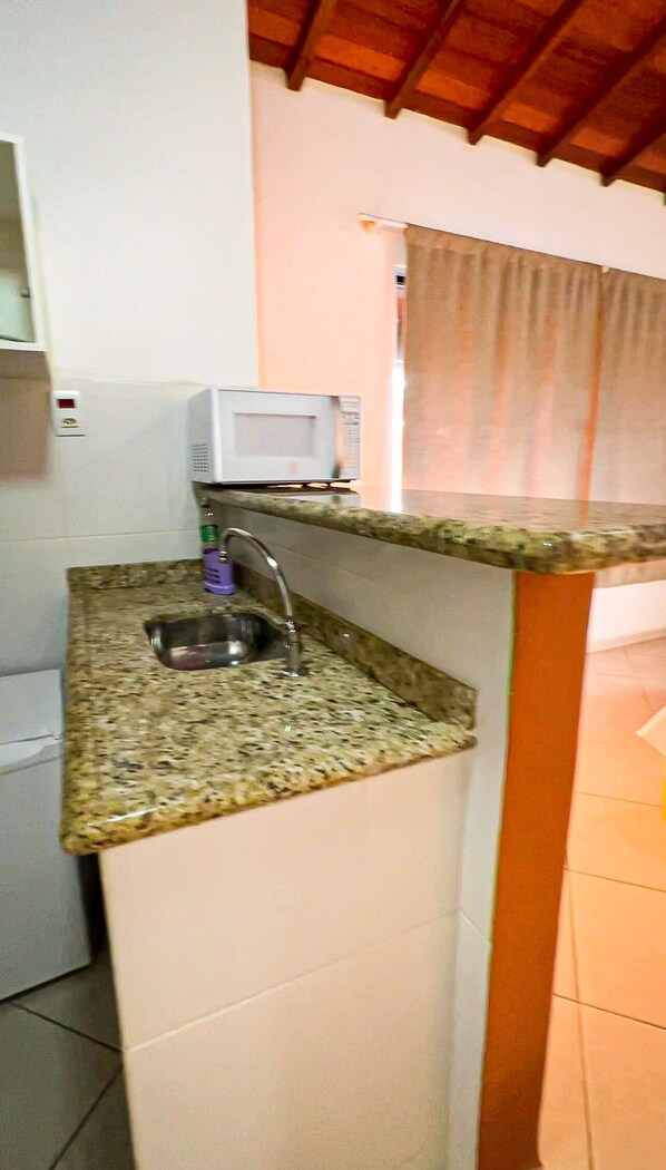 Una pequeña kitchenette con encimera de granito, un fregadero, un microondas y una ventana con cortinas.