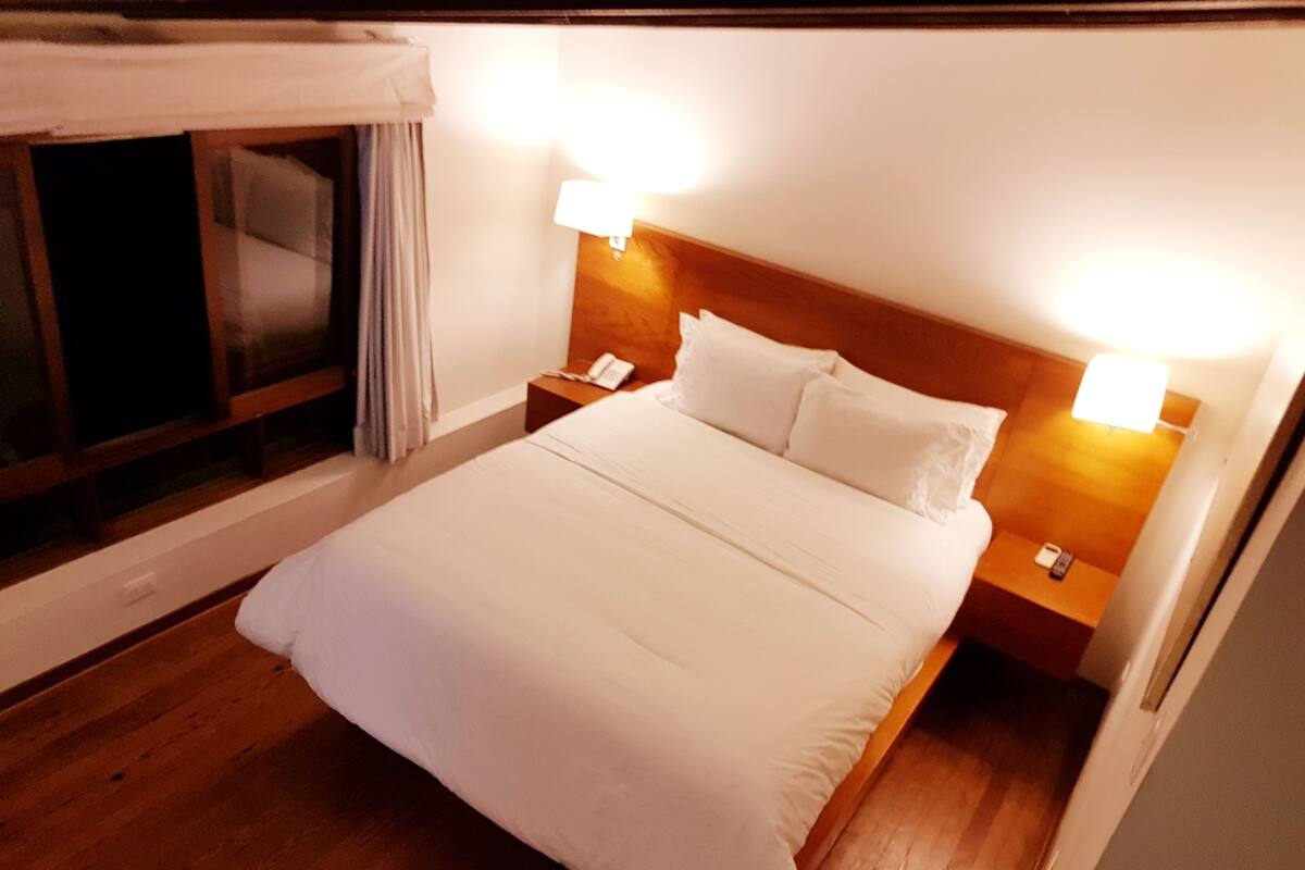 A hotel room with a large bed, two bedside tables with lamps, a window with curtains, and a wooden floor.