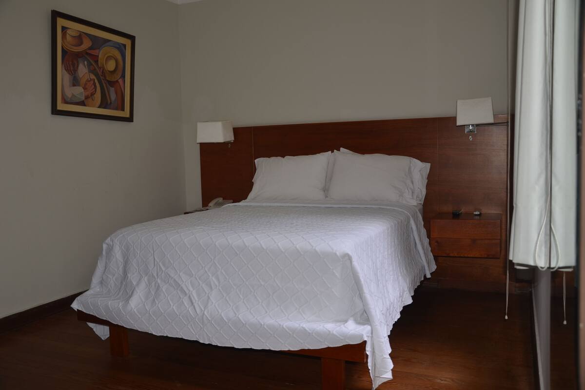 A neatly made bed with a white quilt, a wooden headboard, bedside lamps, and a painting on the wall.