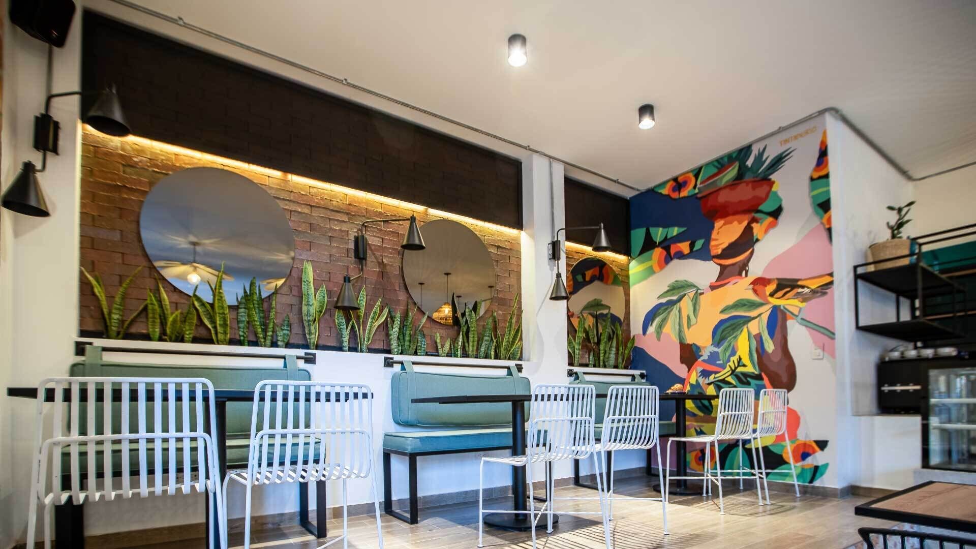 Zona de bar moderna con pared de ladrillo, espejos amplios y un mural con temática tropical. Hay sillas blancas y una barra.