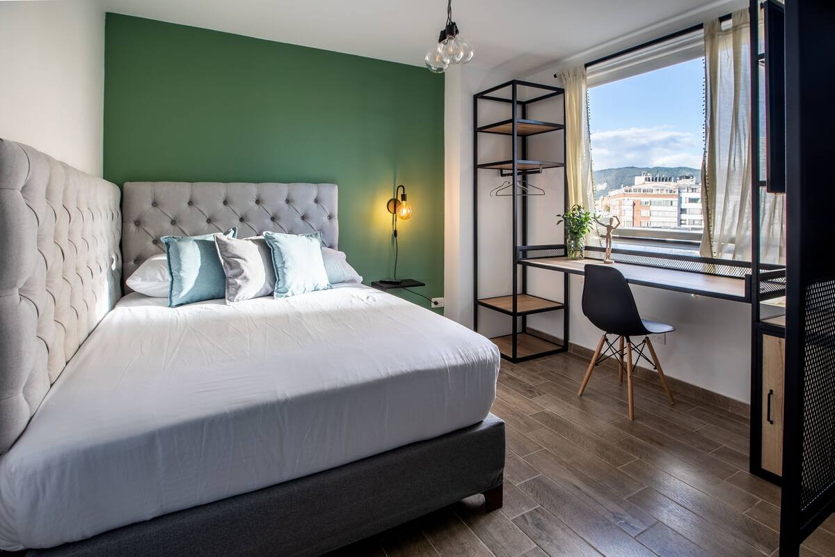 Quarto moderno com cabeceira estofada, uma cama grande, uma escrivaninha com cadeira e vista para a cidade através de uma janela.