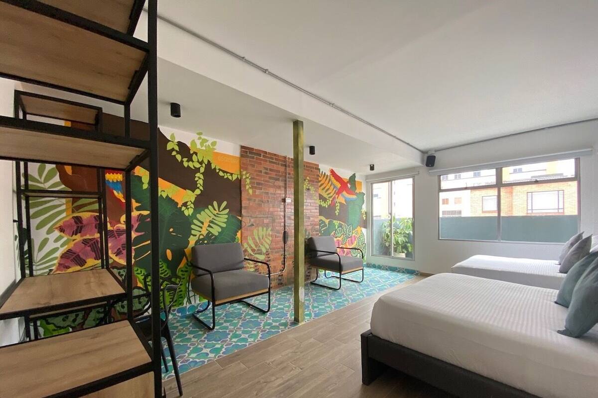 Quarto de hotel moderno com uma cama grande, duas cadeiras, uma mesa pequena e um mural colorido na parede.