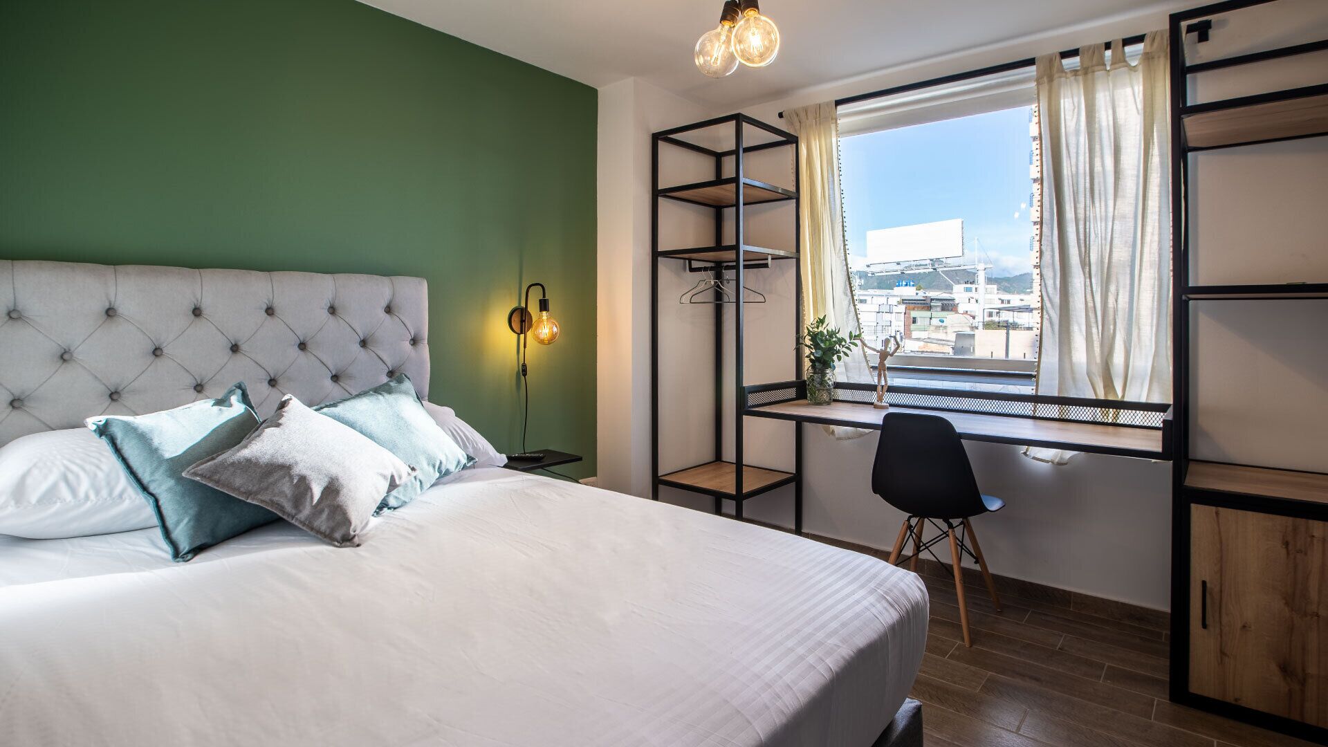 A bedroom with a bed, a desk, a chair, a hanging light fixture, and a window with a view.