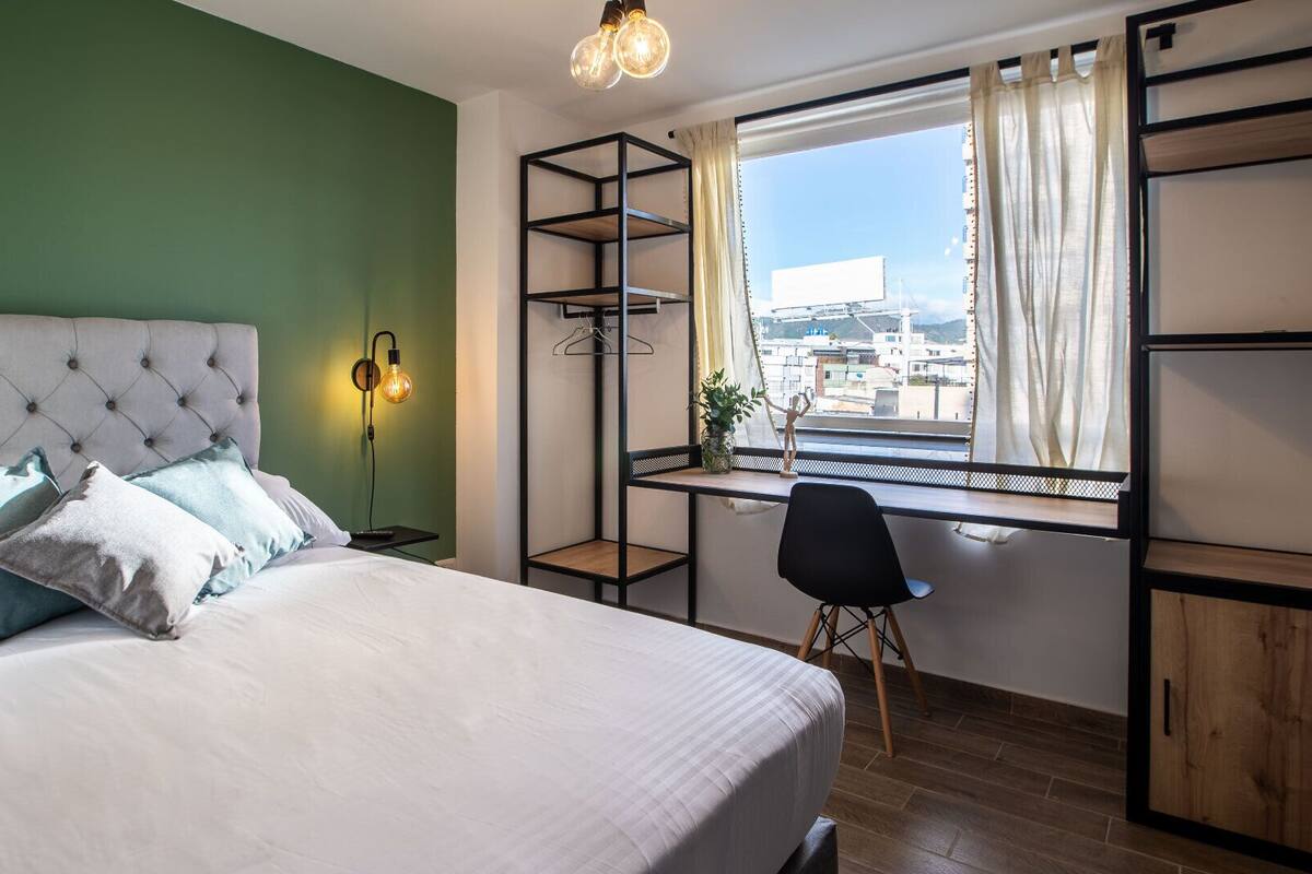 Quarto com cama, escrivaninha, cadeira, luminária pendente e janela com vista.