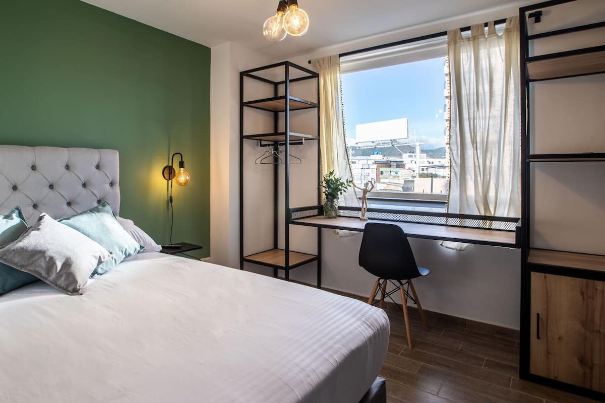 A bedroom with a bed, a desk, a chair, a hanging light fixture, and a window with a view.