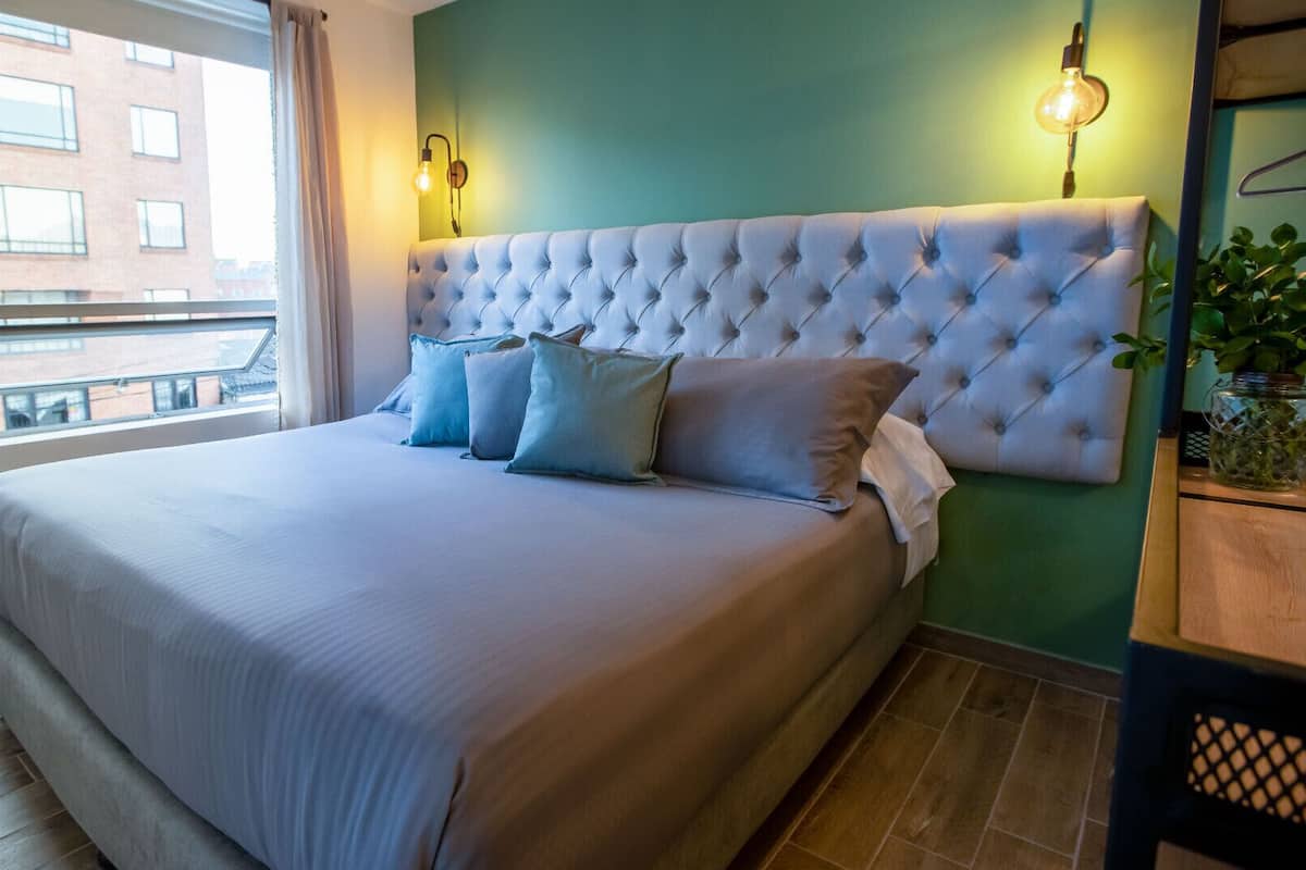 Cama bem arrumada com cabeceira estofada, janela com cortinas e luminária fixada na parede.