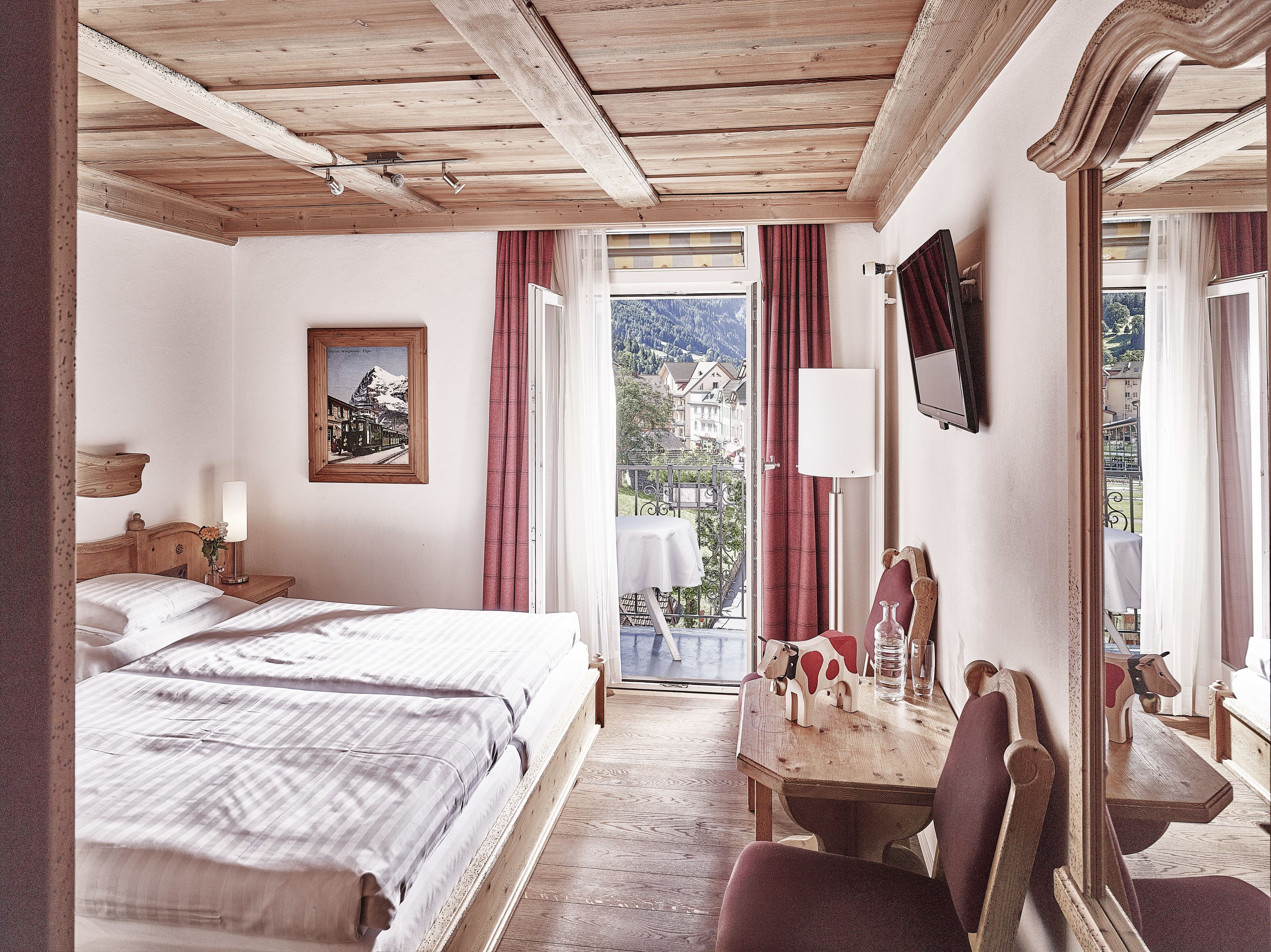A hotel room with a wooden ceiling, a bed, a table with chairs, a mirror, and a view of a mountain and buildings through the window.