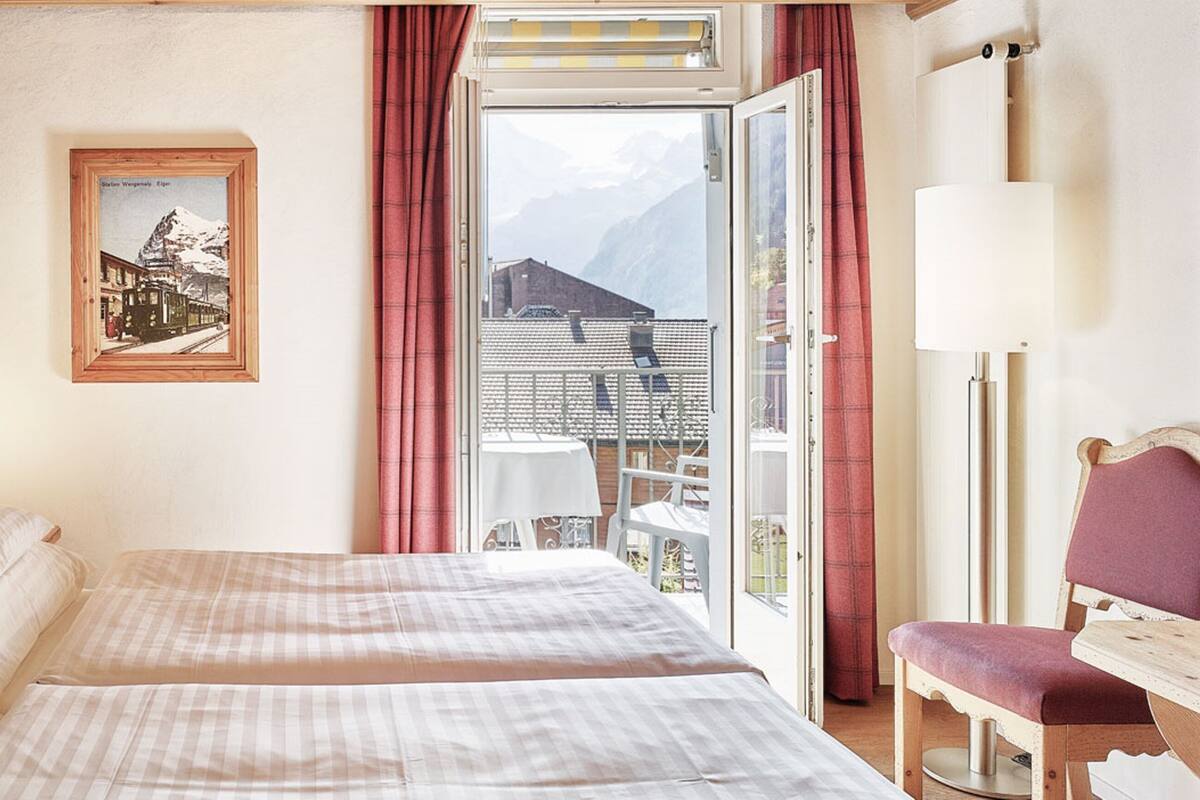 Een hotelkamer met een bed, een stoel, een tafel, een schilderij aan de muur en uitzicht op bergen door het raam.