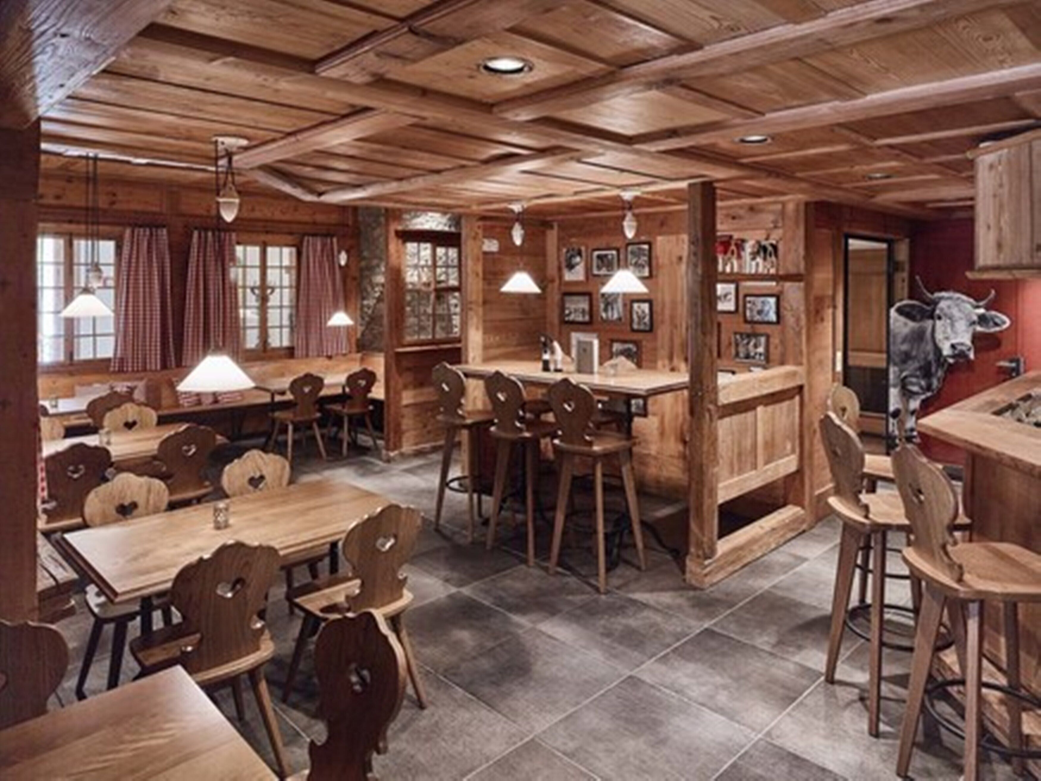 Ein rustikales Interieur aus Holz mit Tischen, Stühlen und einer Bar. An der Wand hängt eine Kuh als Dekoration.