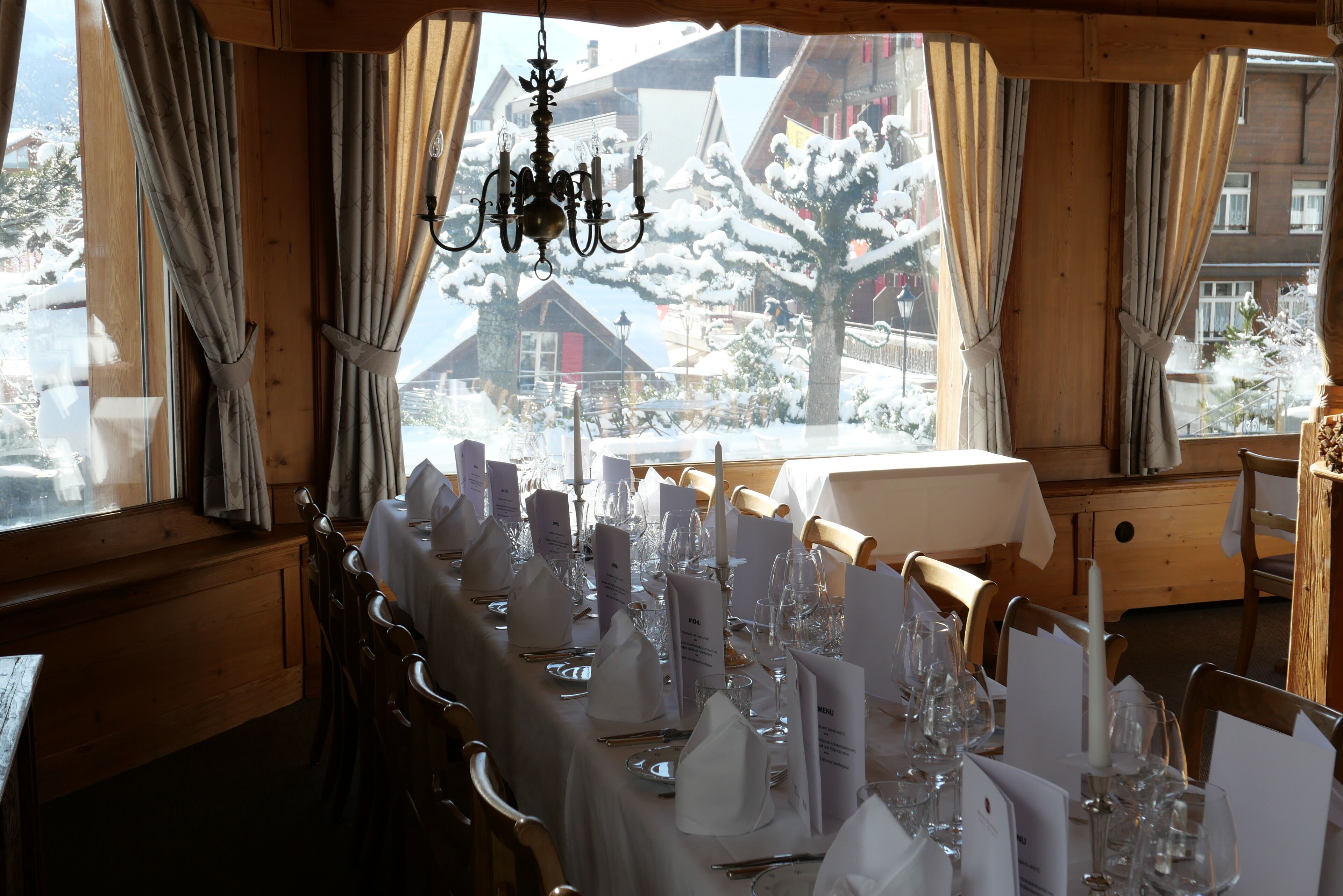 Uma sala de jantar com mesa posta para refeição, um lustre e vista para uma aldeia coberta de neve através de amplas janelas.