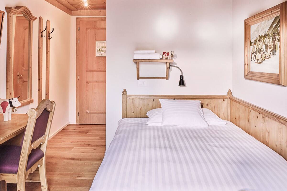 Een houten slaapkamer met een bed, een stoel, een houten bureau en een plank met handdoeken.
