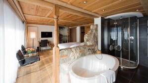A modern bathroom with a stone accent wall, a freestanding bathtub, and a glass-enclosed shower. - Hotel Schönegg Wengen (Lauterbrunnen)