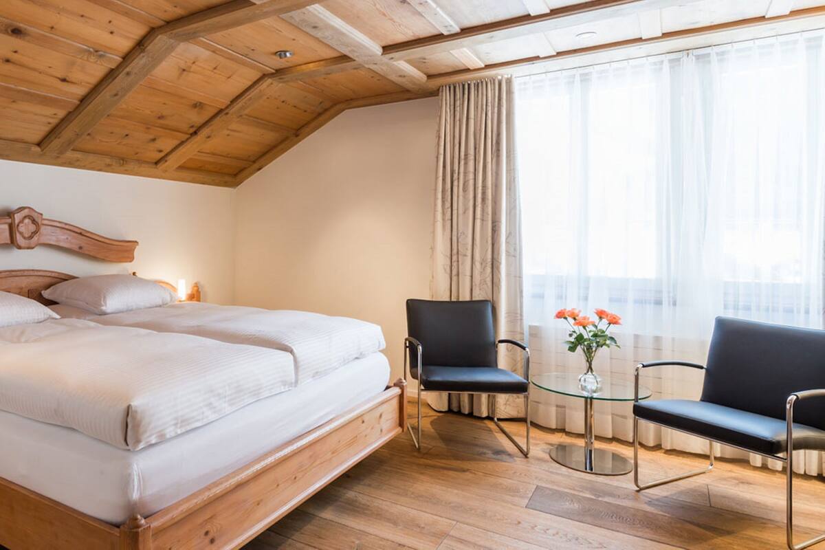Een slaapkamer met houten frame, met een groot bed, twee stoelen, een klein tafeltje en een nachtkastje.