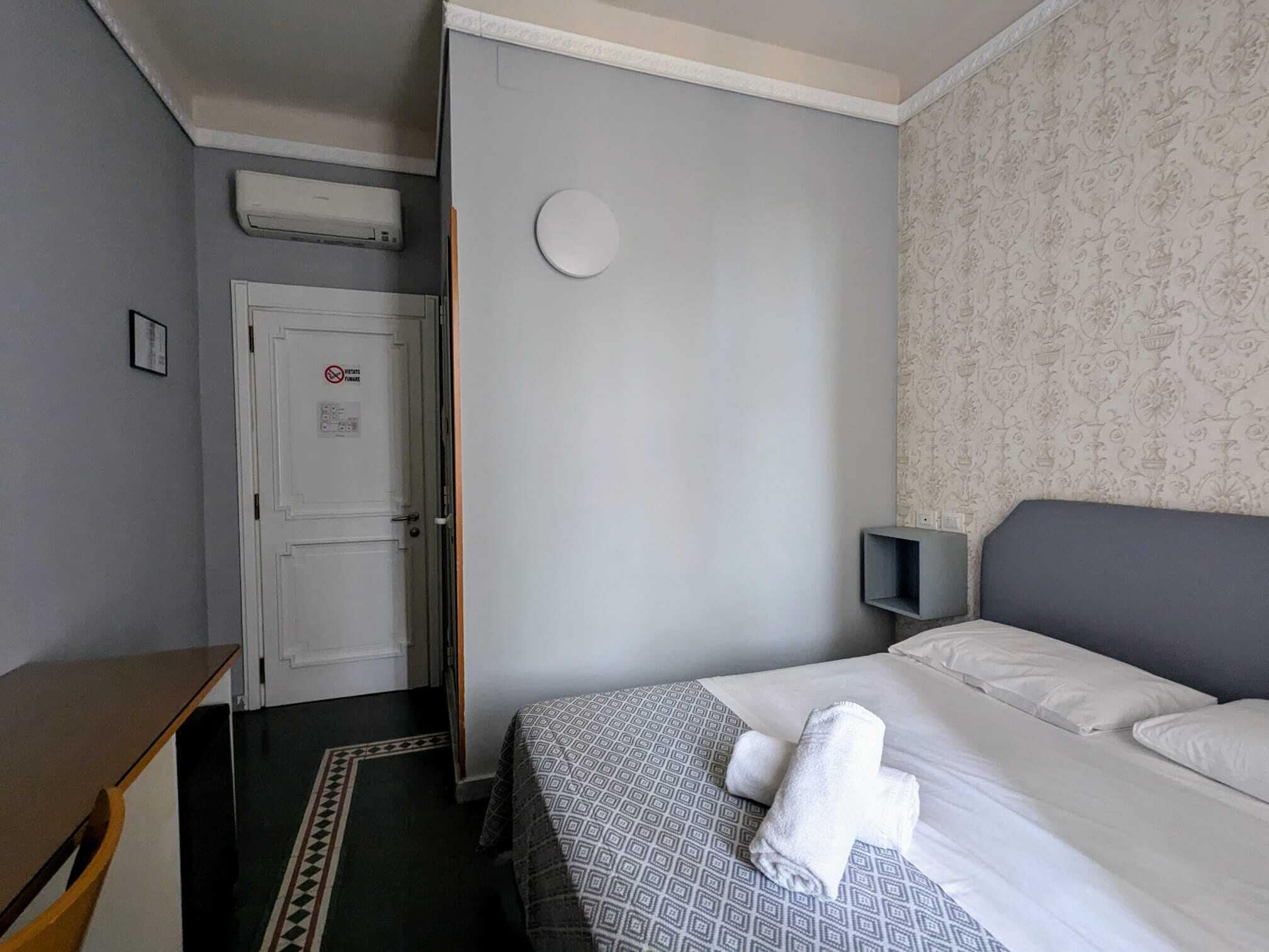 A hotel room with a bed, a desk, a chair, a patterned wall, and a door with a no smoking sign.