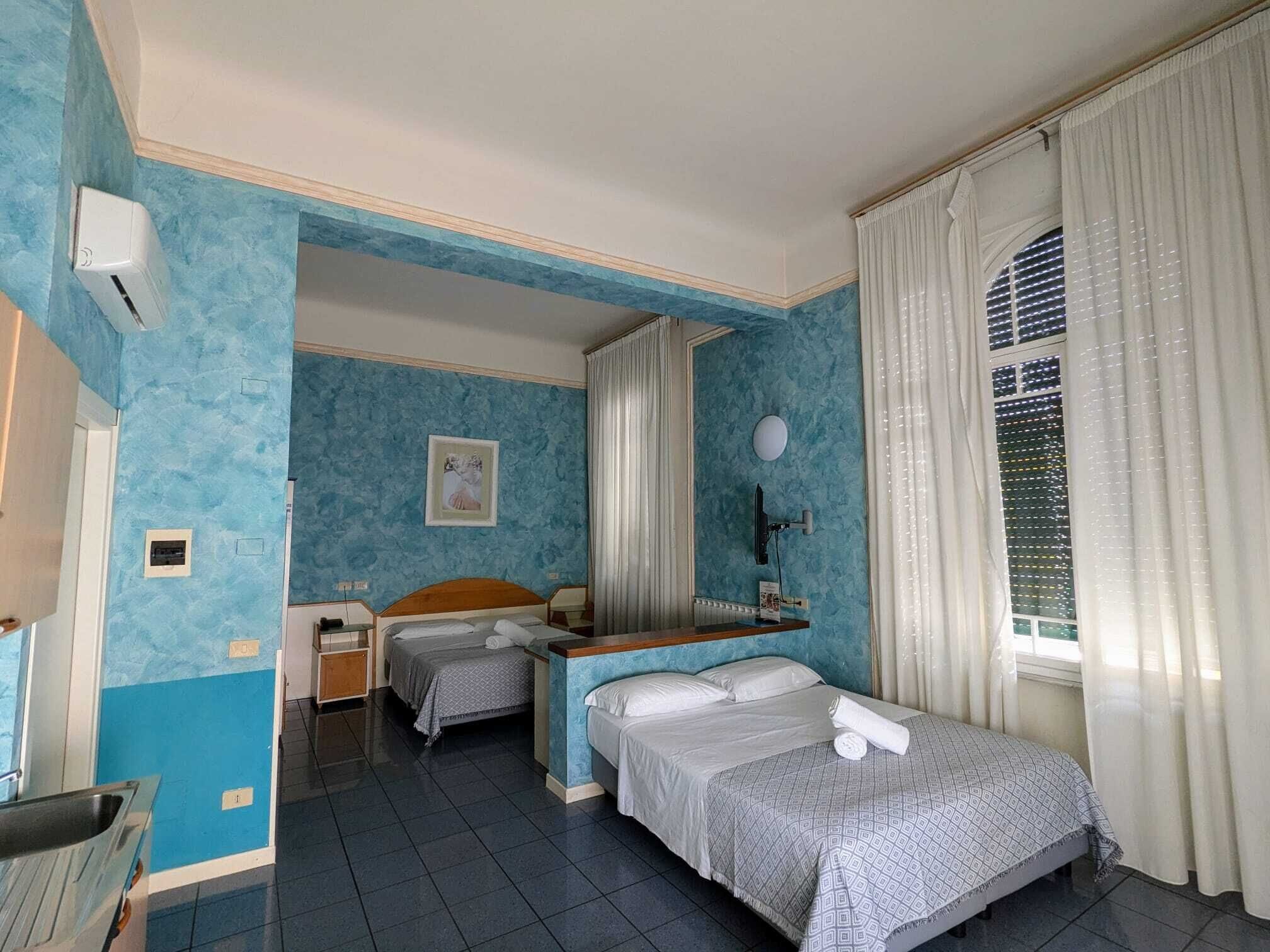 A hotel room with two beds, a desk, a chair, a window with curtains, and a wall-mounted air conditioner.