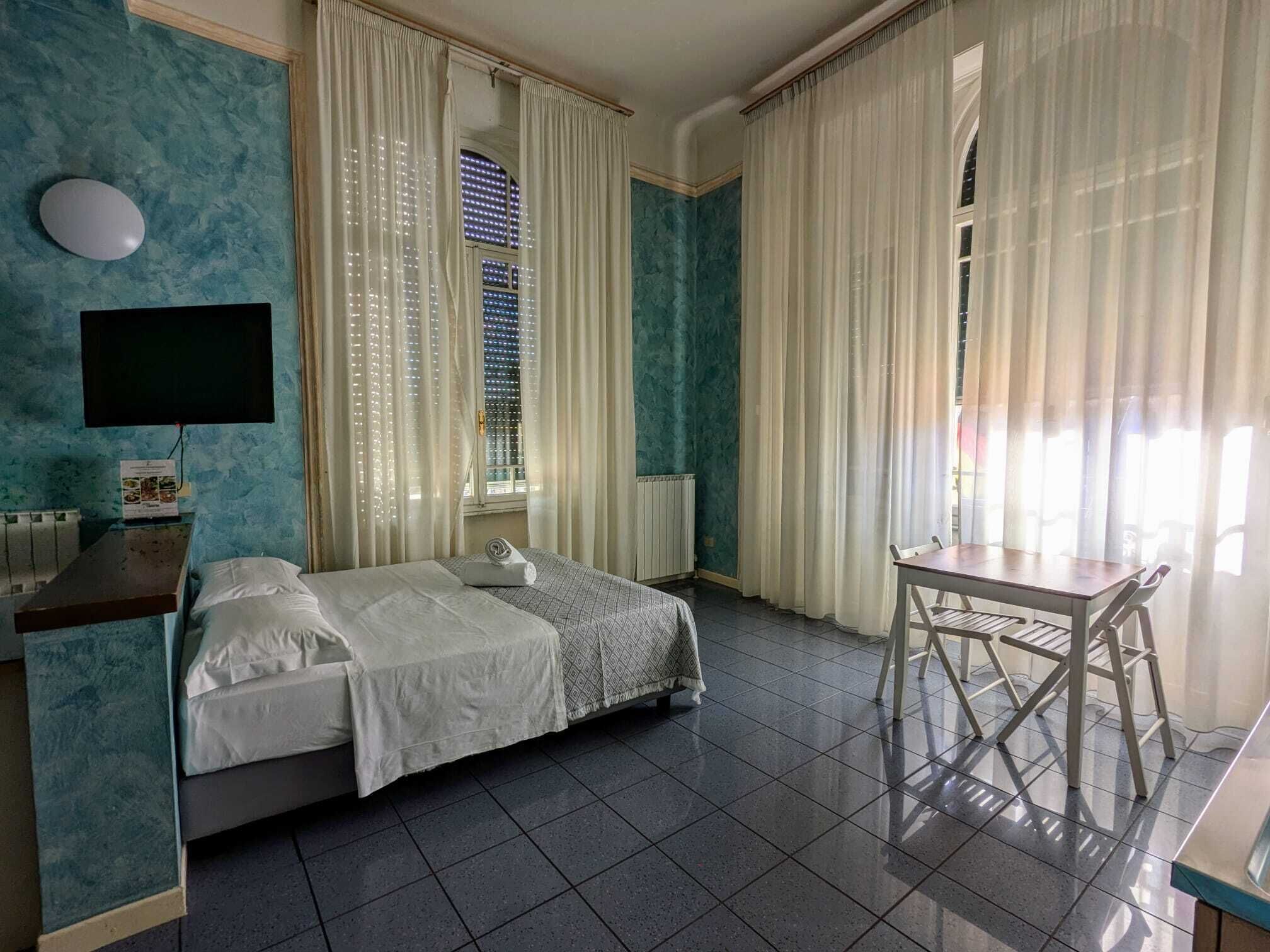 Habitación de hotel con cama, escritorio y silla. Se ve una ventana con cortinas, un televisor de pantalla plana en la pared y un radiador.