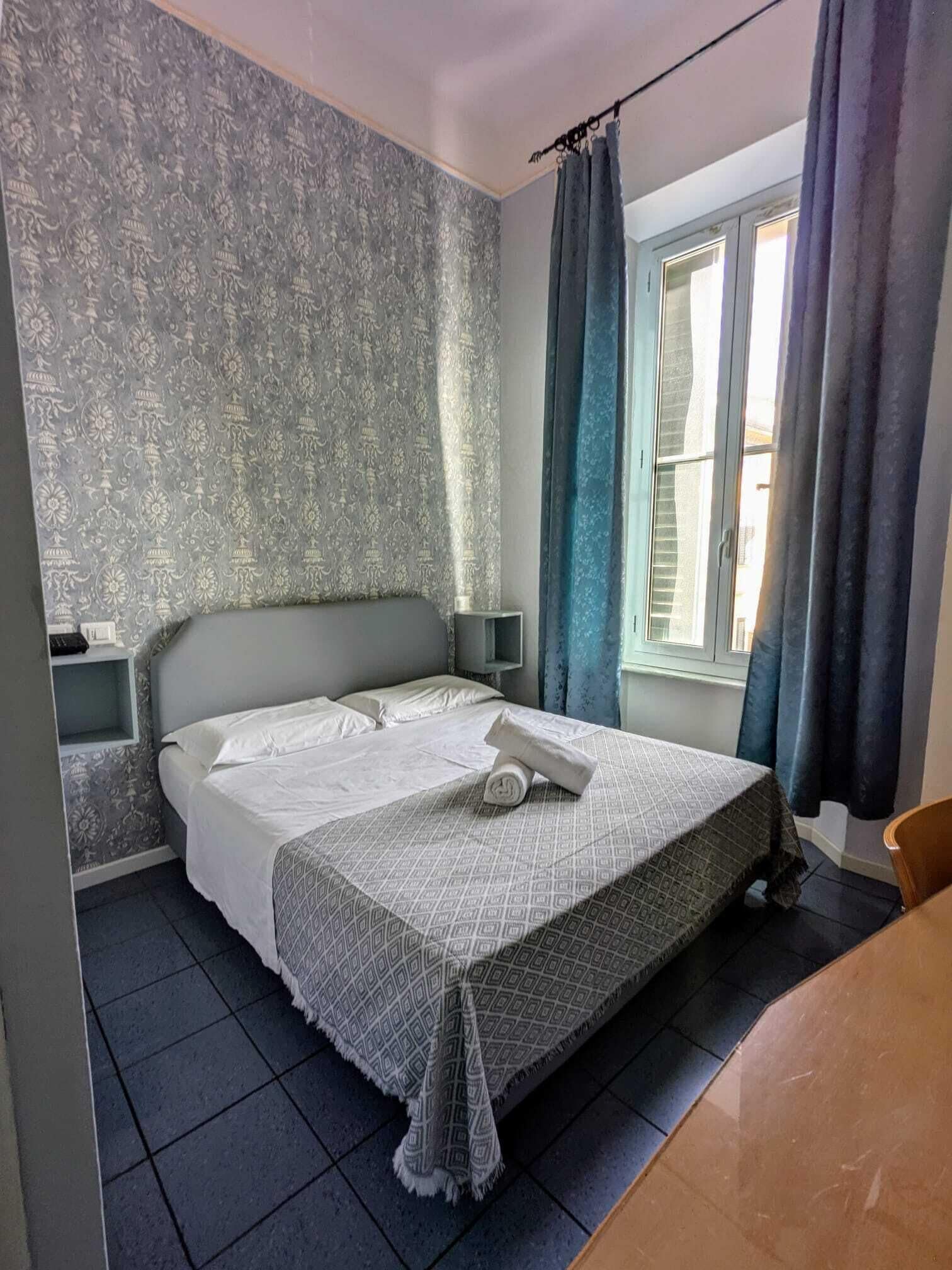 A bedroom with a bed, nightstands, a window with curtains, and a tiled floor.