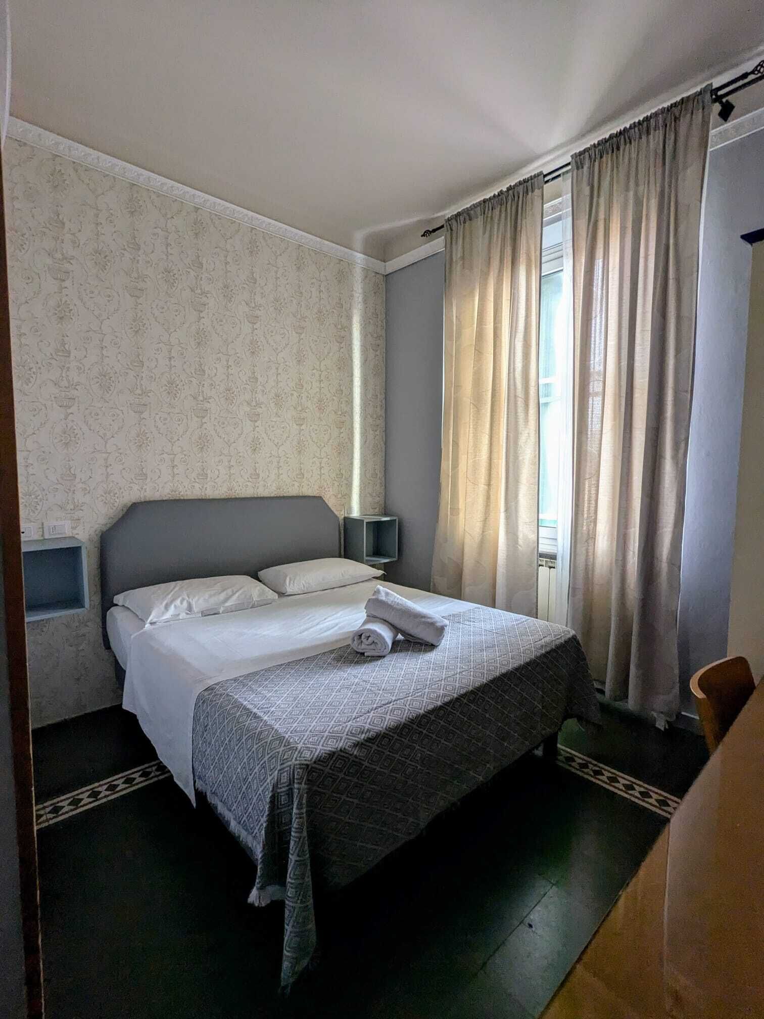 Habitación de hotel ordenada con cama, mesita de noche, lámpara y ventana con cortinas.
