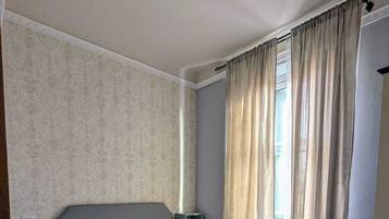 Ein ordentlich eingerichtetes Hotelzimmer mit Bett, Nachttisch, Lampe und einem Fenster mit Vorhängen.