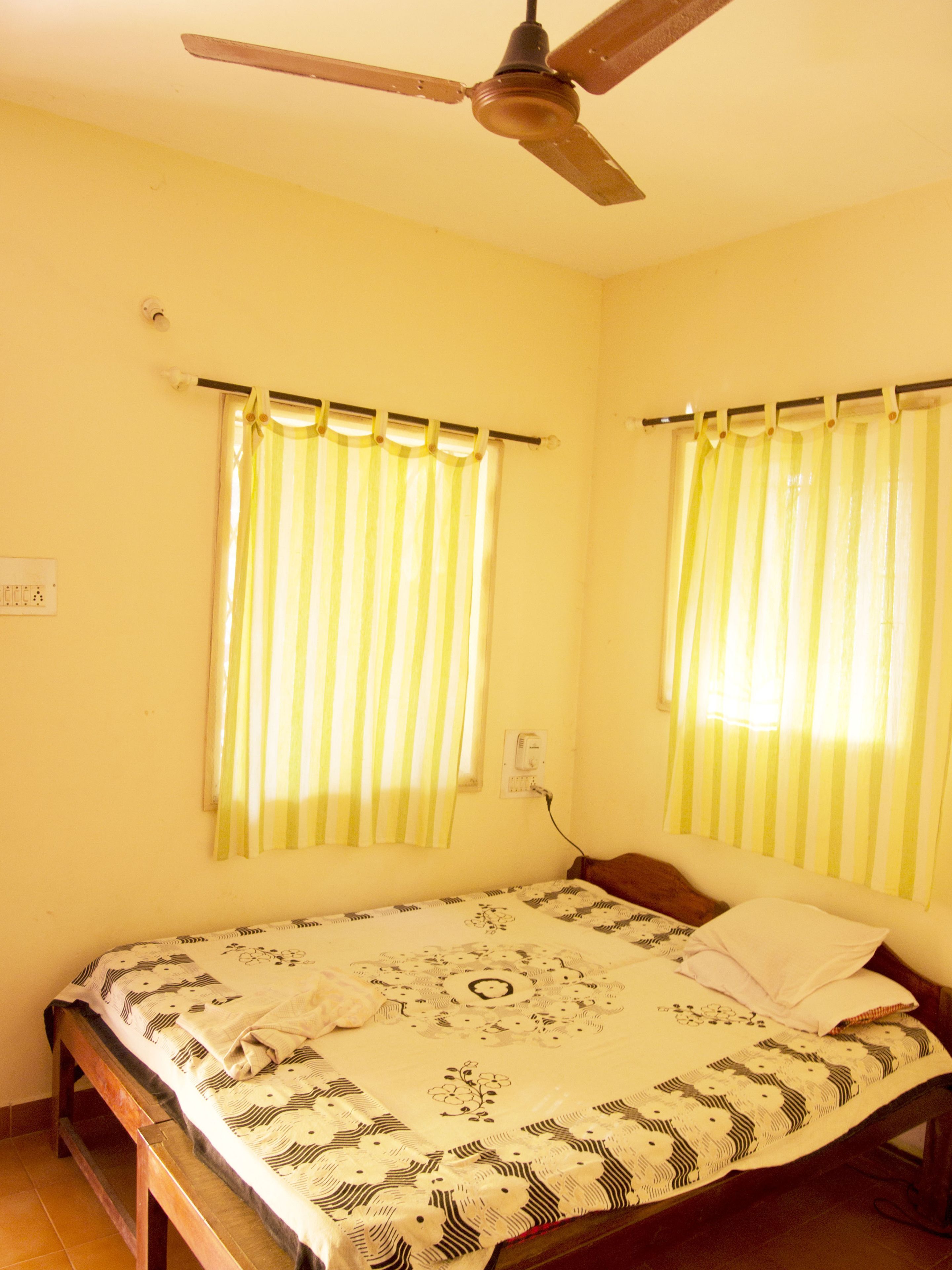 Quarto com cama, ventoinha no teto e duas janelas com cortinas.