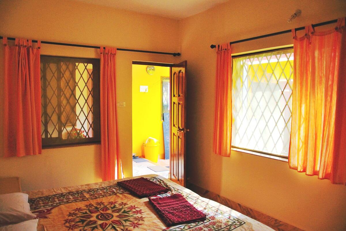 Une pièce avec un lit, deux fenêtres avec des rideaux, une porte donnant sur une autre pièce et un mur jaune.