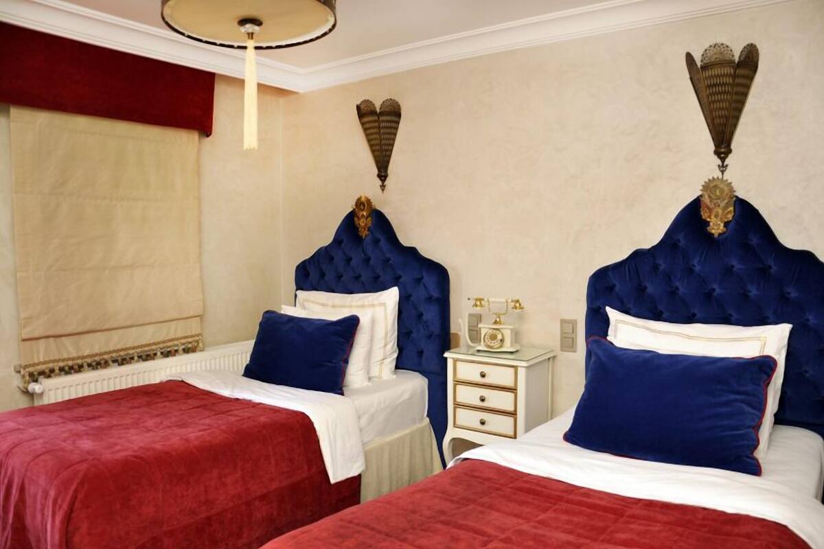 Quarto de hotel com duas camas, cada uma com cabeceira azul e roupa de cama vermelha. Há uma mesa de cabeceira branca com um rádio entre as camas.